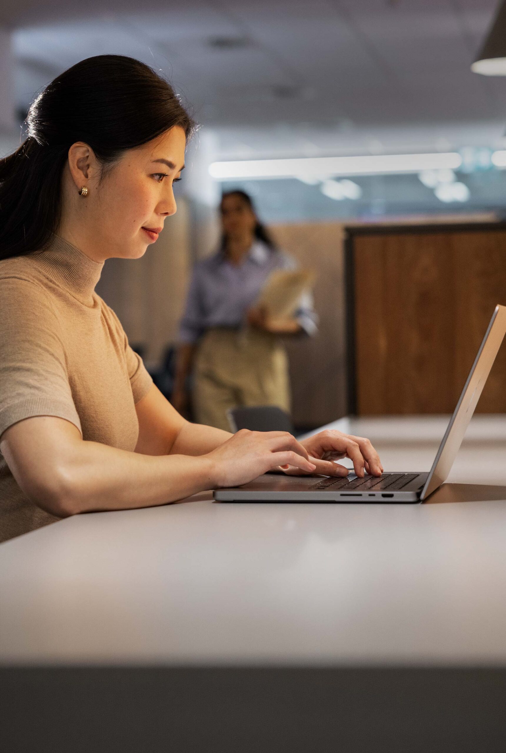 A woman works in an office environment, typing on her laptop. There are more people walking and working in the background.