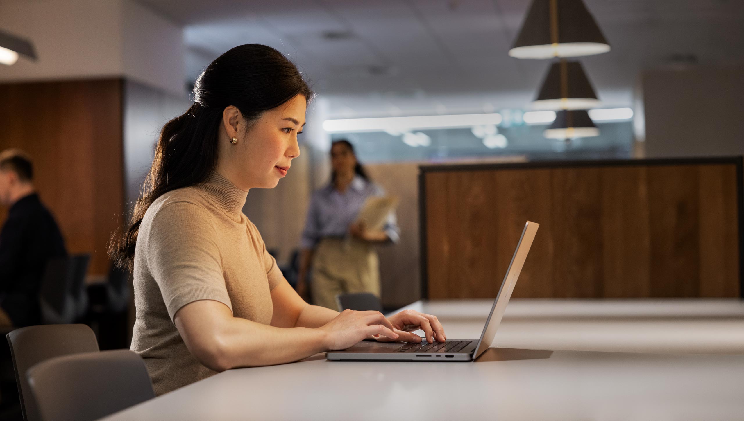 A woman works in an office environment, typing on her laptop. There are more people walking and working in the background.