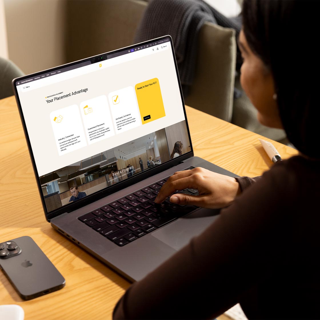 A young woman looks at her laptop sitting at a desk. The screen shows a webpage.
