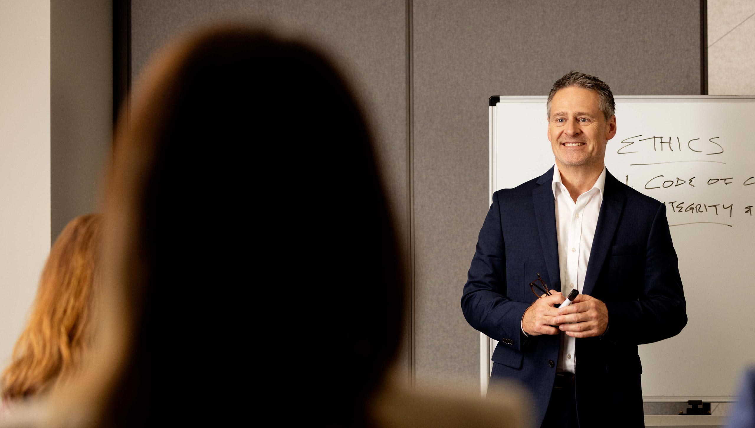 A male teacher stands infront of a whiteboard which says 'Ethics'. Students sit in front of him, blurred.