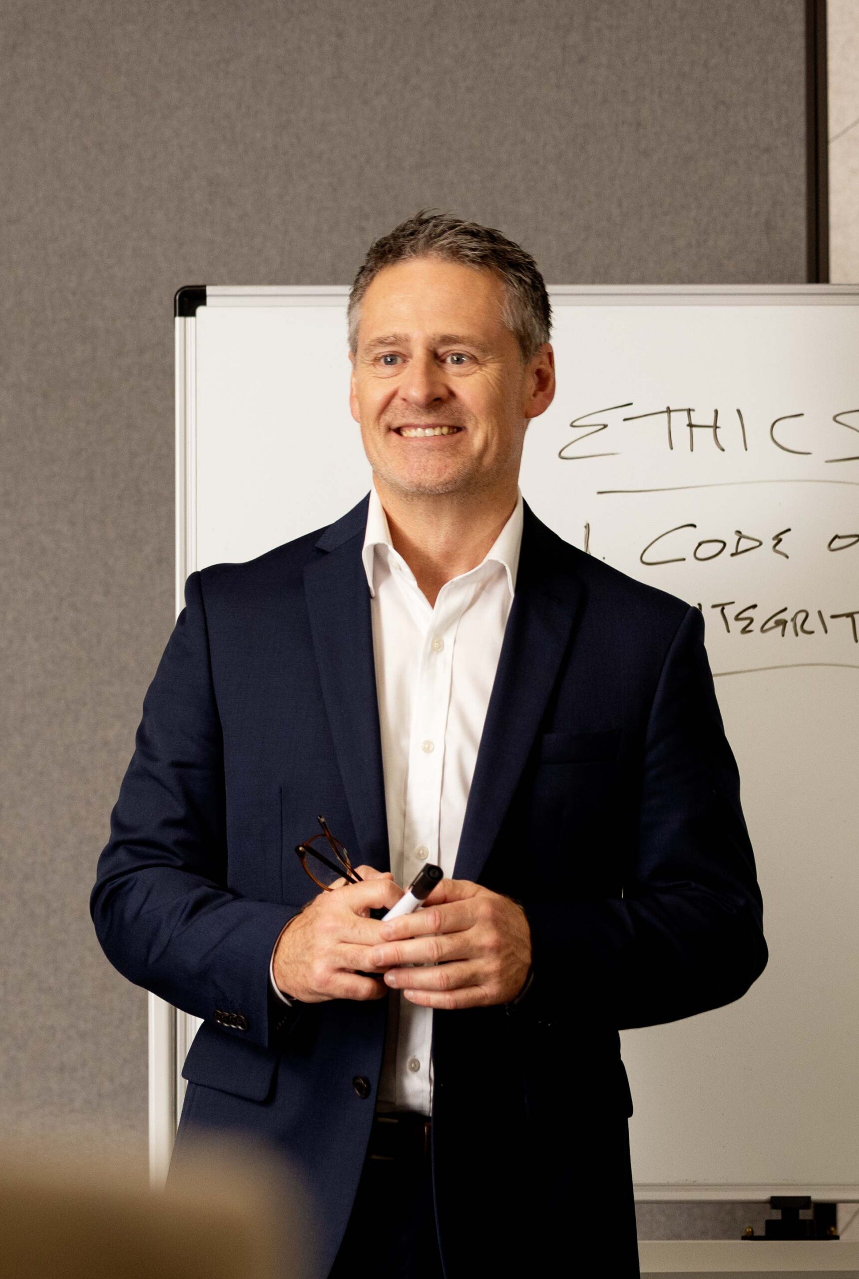 A male teacher stands infront of a whiteboard which says 'Ethics'. Students sit in front of him, blurred.