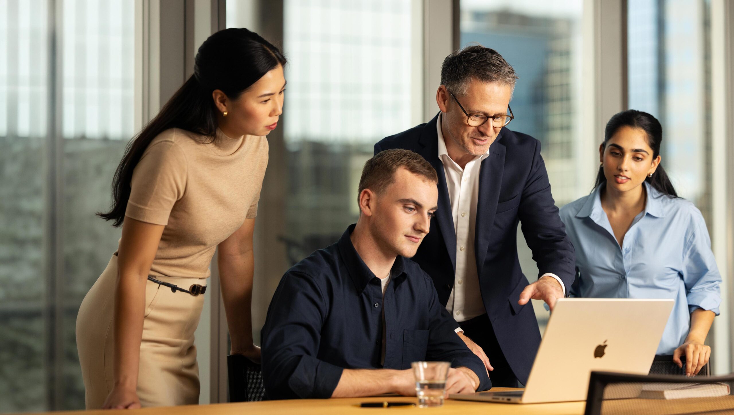 Three people stand around a a fourth person who sits at a table. They are looking at a laptop, while one of the three standing people explains something on the screen.