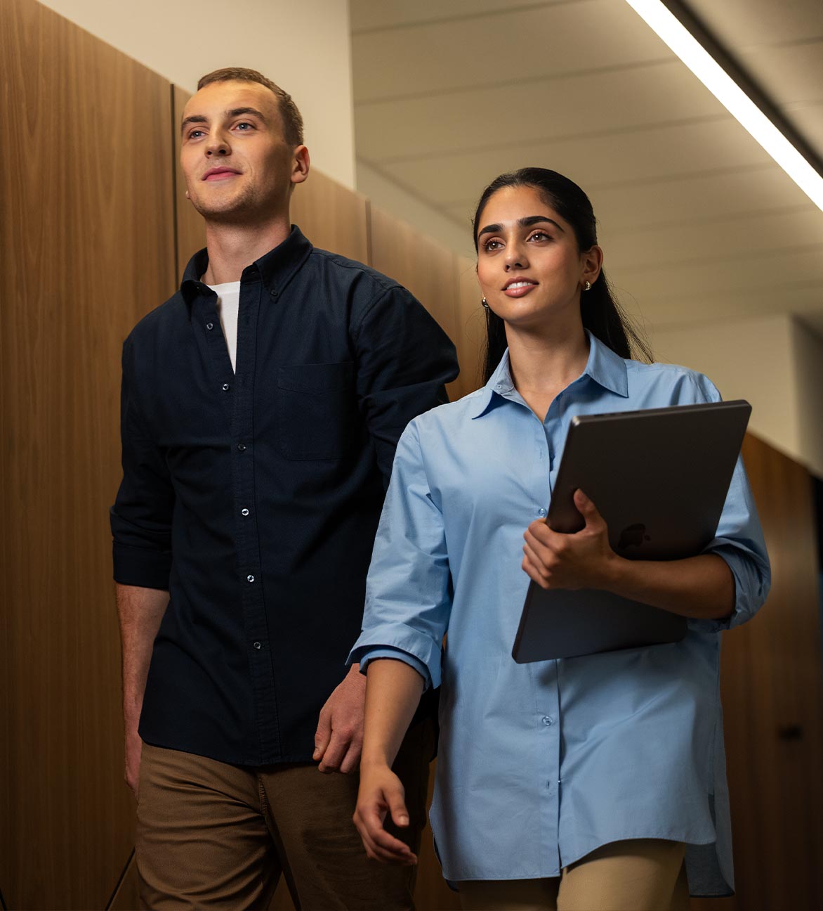 A young man and woman dressed in business casual walk down a hallway with purpose. The woman holds a laptop.
