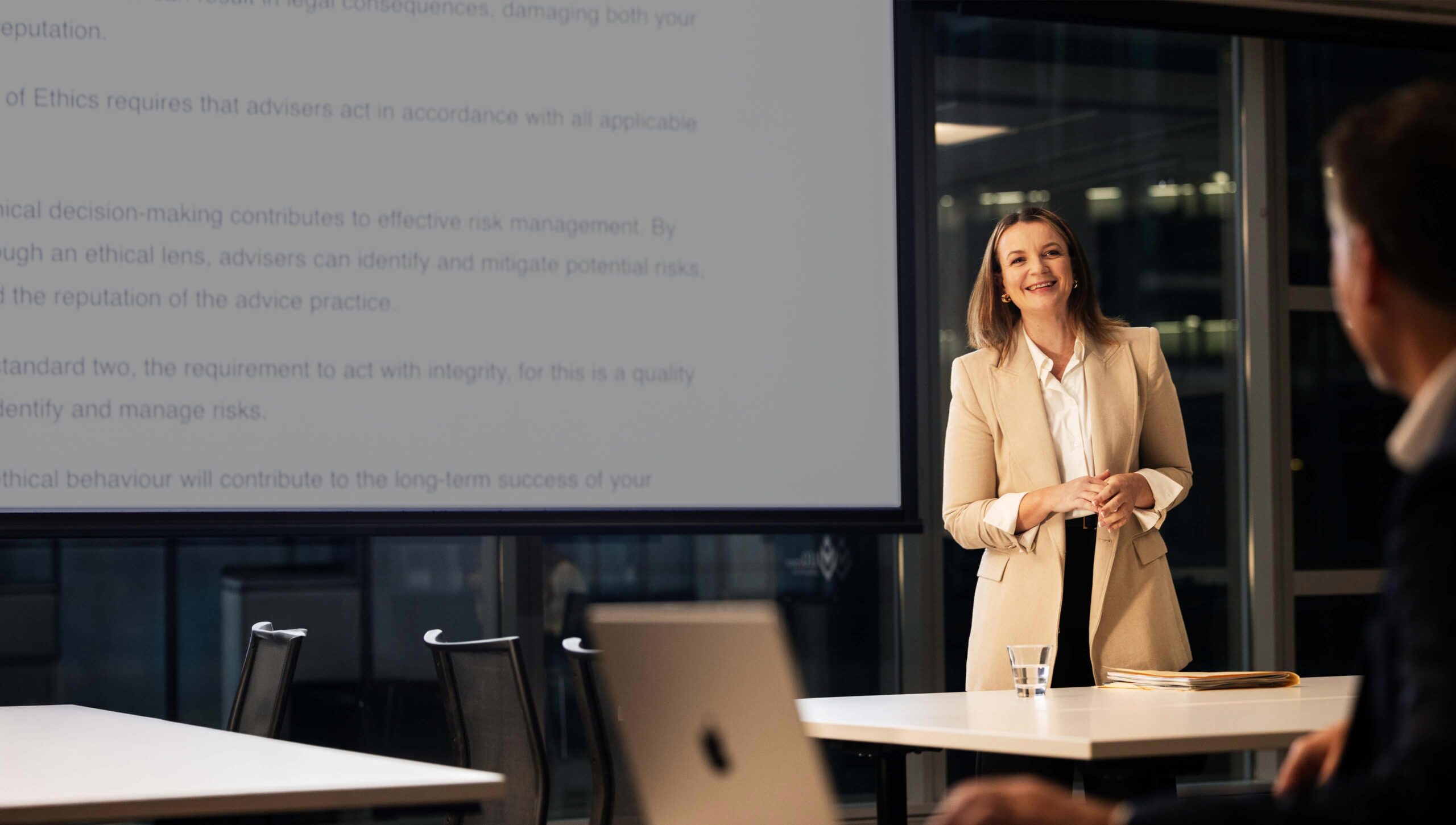 A middle-aged woman stands in front of a projector screen giving a lecture.
